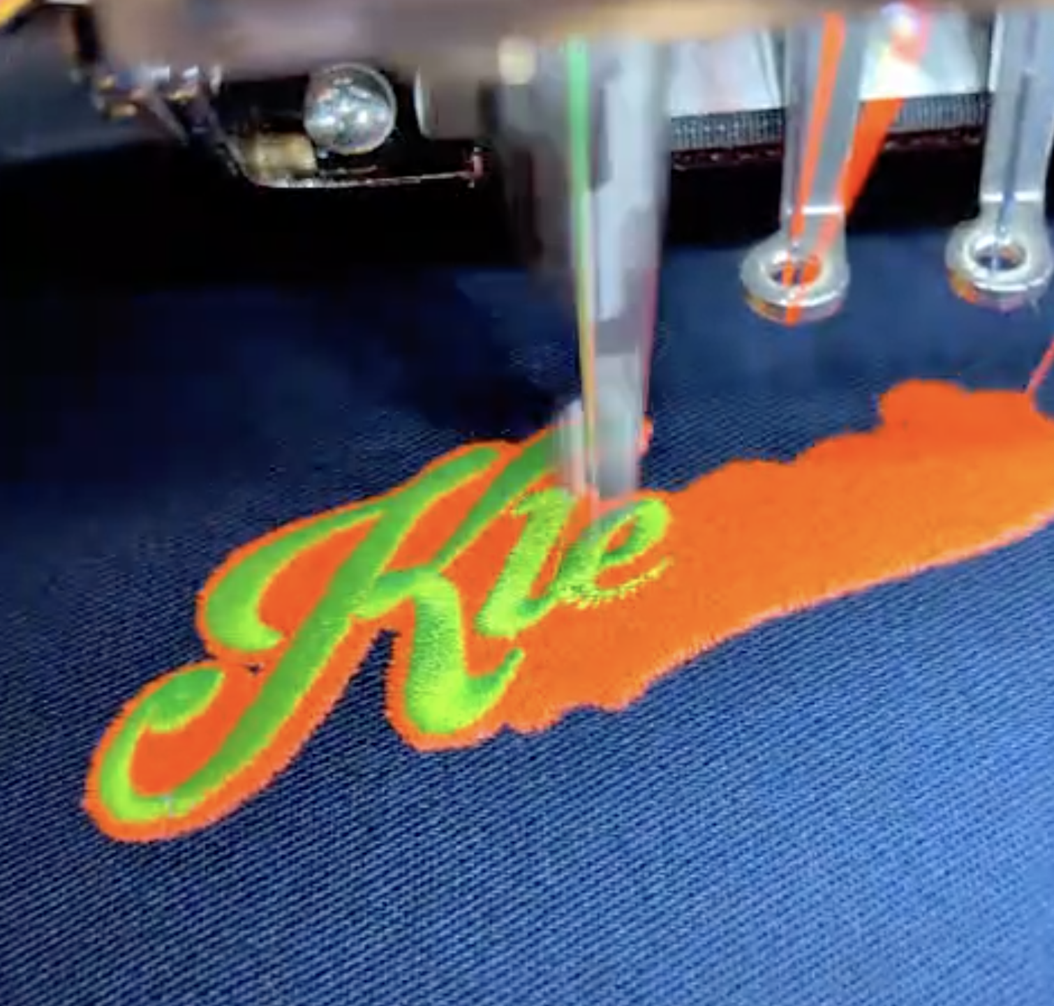 Stickmaschine stickt "Kle" in Grün mit orangefarbener Umrandung auf dunkelblauem Stoff.