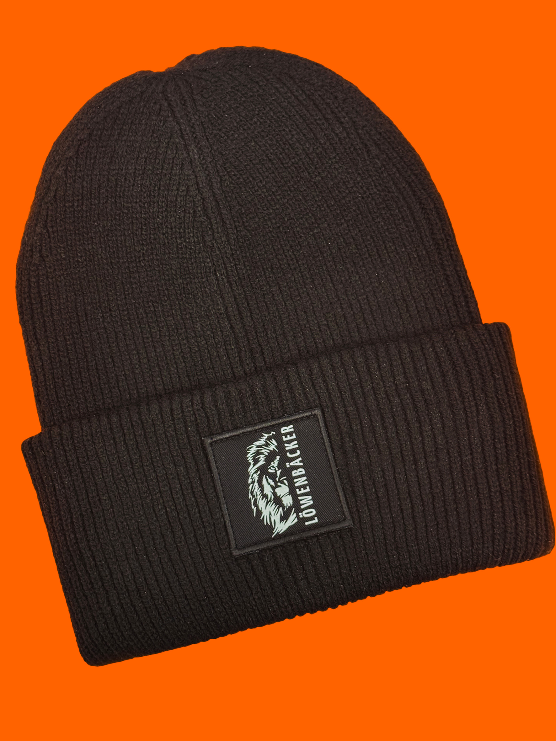 Eine schwarze Strickmütze mit aufgenähtem Patch und Löwenmotiv auf orangem Hintergrund