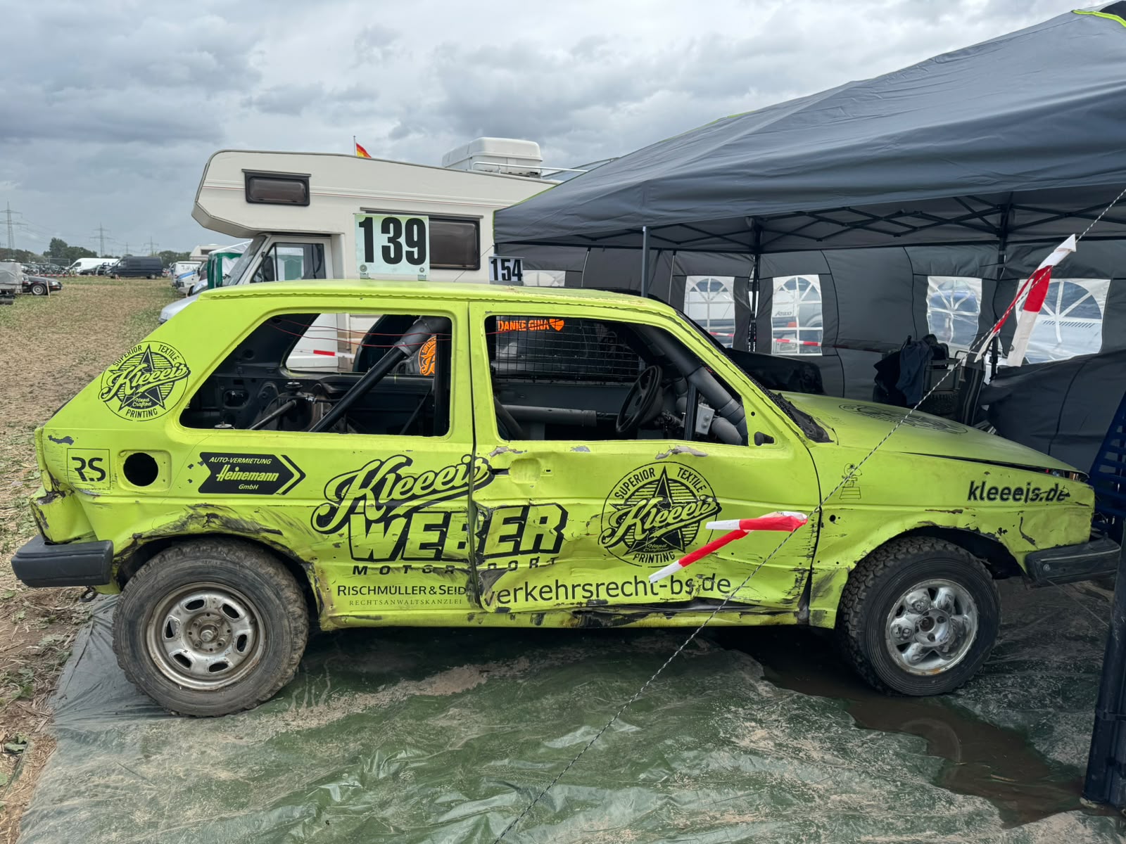 Ein knallgrunes verbeultes Stockcar Rennauto steht vor einem Wohnmobil