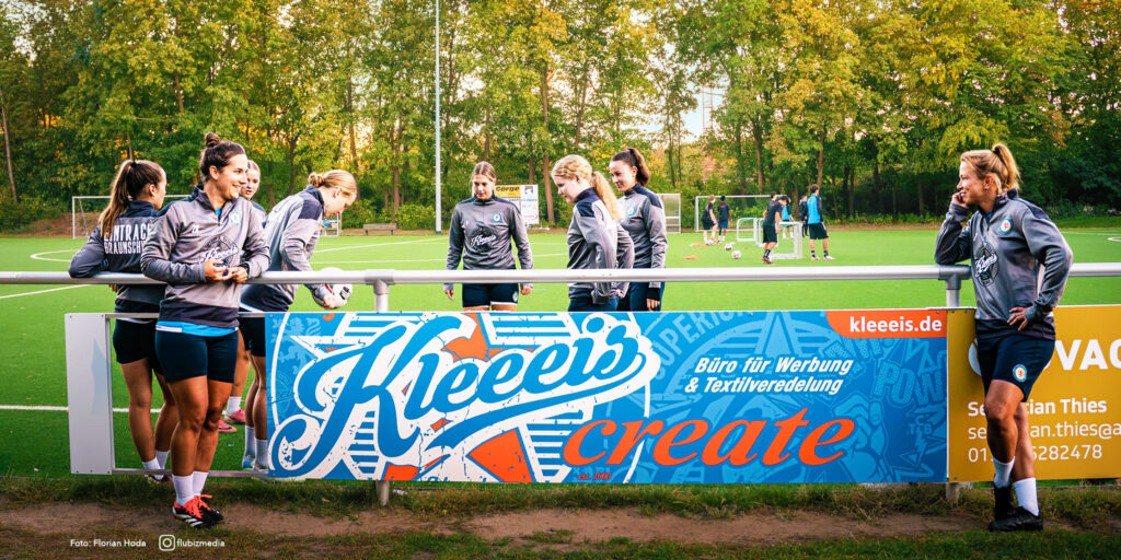 Fussballerinnen in grauen Trikots stehen entspannt an einer Bande mit Werbebanner