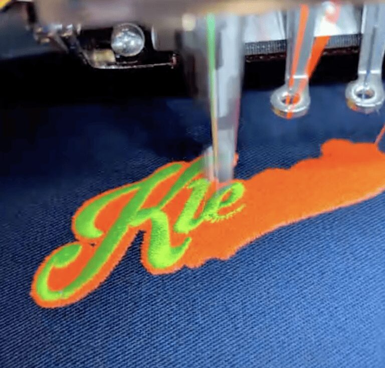 Stickmaschine bestickt blauen Stoff mit gruenem Schriftzug auf Orange